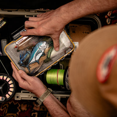 A close-up lifestyle shot of a person holding a rectangular fly fishing box , including a GT Giant Trevally Sticker and spools of line, inside an open storage case. With Fly Rod.