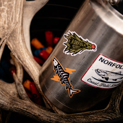 A stainless steel water bottle with fly fishing green woolly bugger stickers, topped with a tan Norfolk Wild cap, set on a rustic wooden surface with hunting antlers in the background.