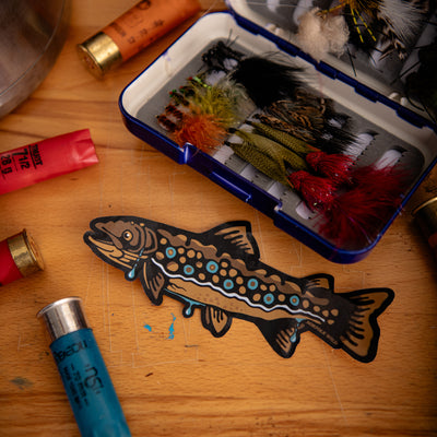 A brown trout sticker or decal with blue and orange spots and black outlines lies on a wooden table, with a blue fly box containing fishing lures and shotgun shells nearby.