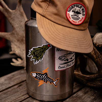 A stainless steel water bottle with fly fishing green woolly bugger stickers, topped with a tan Norfolk Wild cap, set on a rustic wooden surface with hunting antlers in the background.