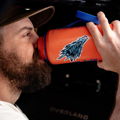 A close-up lifestyle shot of a man drinking from an orange nalgene water bottle with a blue cap and a blue 'Norfolk State' woolly bugger fly fishing sticker. The background is a dark vehicle interior.